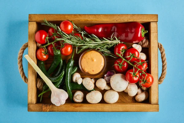 Вид на вкусный соус в миске рядом со свежими зрелыми овощами, розмарином и грибами в деревянной коробке на синем фоне — стоковое фото