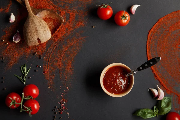 Vista superior de pasta de tomate em tigela perto de tomates cereja, dentes de alho, páprica em pó, alecrim e folhas de manjericão em preto — Fotografia de Stock