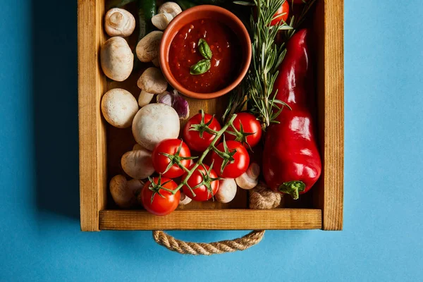 Ansicht von Tomatensauce in Schüssel auf Champignons in der Nähe von roten Kirschtomaten, Rosmarin und Chili in Holzkiste auf blau — Stockfoto
