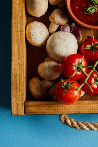 Top view of tomato sauce in bowl near mushrooms, red cherry tomatoes and garlic cloves in wooden box on blue — Stock Photo