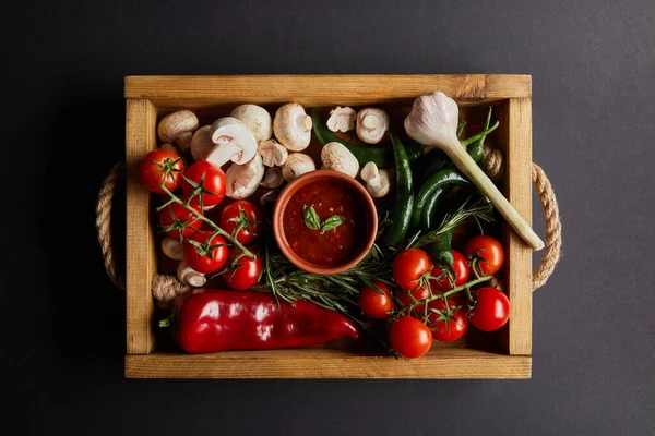 Ansicht von Tomatensauce mit Basilikumblättern in der Nähe von Kirschtomaten, grünen Chilischoten, Pilzen und Rosmarin in Holzkiste auf schwarz — Stockfoto