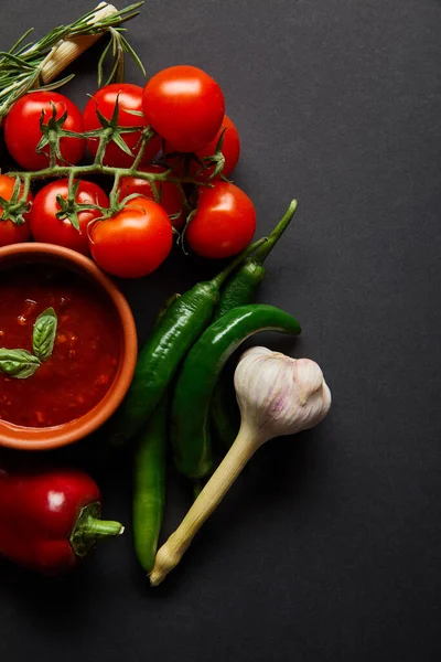 Top view of organic cherry tomatoes, garlic, tomato sauce in bowl and rosemary near chili peppers on black — Stock Photo