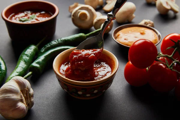 Selektiver Fokus des Löffels in leckerer Tomatensauce nahe reifem Gemüse auf schwarzem — Stockfoto