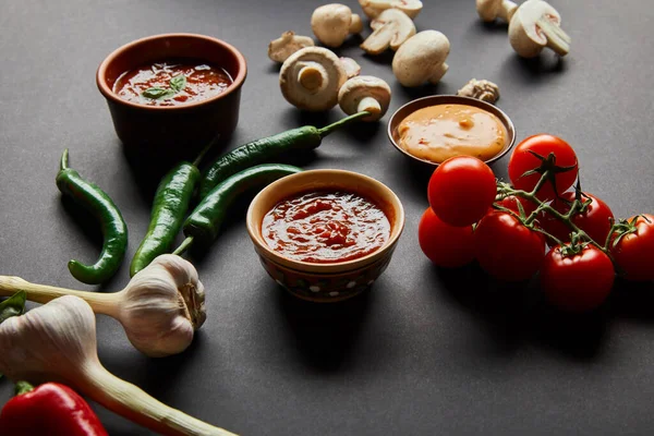 Selective focus of tasty sauces in bowls near ripe cherry tomatoes, green chili peppers, mushrooms and garlic on black — Stock Photo