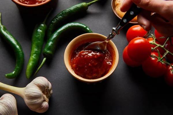 Vista cortada de uma pessoa segurando colher perto de molho de tomate saboroso e legumes em preto — Fotografia de Stock