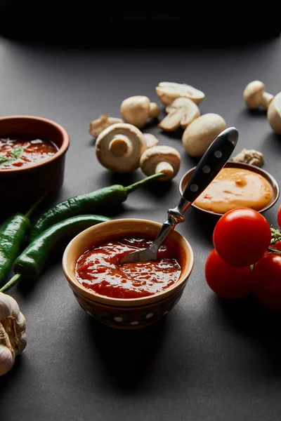 Selective focus of tasty sauces in bowls near ripe cherry tomatoes, green chili peppers and mushrooms on black — Stock Photo