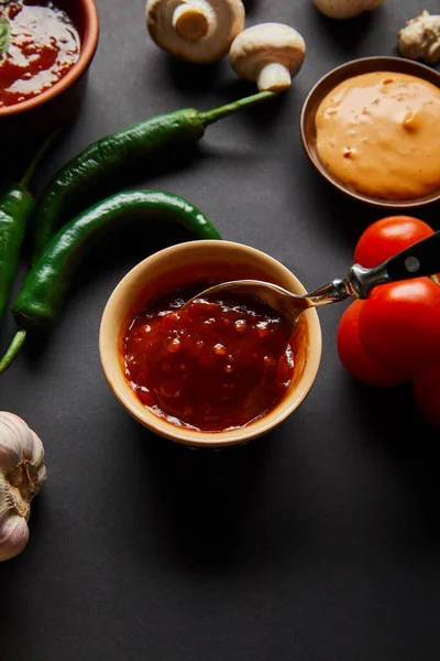 Top view of tasty sauces in bowls near ripe cherry tomatoes, green chili peppers and mushrooms on black — Stock Photo