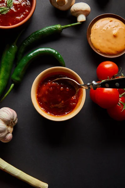 Top view of tasty sauces in bowls near ripe cherry tomatoes, garlic, green chili peppers and mushrooms on black — Stock Photo