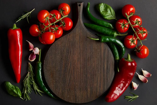 Top view of wooden chopping board near ripe cherry tomatoes, garlic cloves, rosemary, basil leaves and green chili peppers on black — Stock Photo