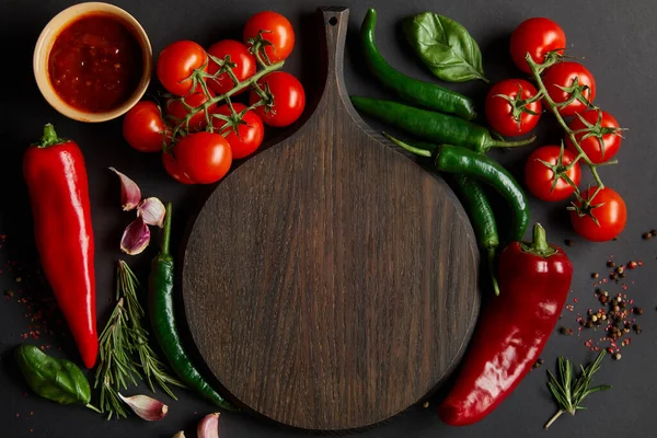 Top view of chopping board near ripe cherry tomatoes, garlic cloves, rosemary, peppercorns, basil leaves and green chili peppers on black — Stock Photo