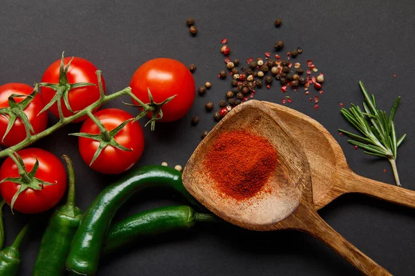 Vue de dessus de tomates cerises rouges, romarin, grains de poivre, cuillères en bois avec poudre de paprika et piments verts sur noir — Photo de stock