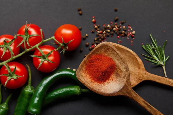 Vista dall'alto di pomodorini rossi ciliegini, rosmarino, grani di pepe, cucchiai di legno con polvere di paprika e peperoncino verde su nero — Foto stock