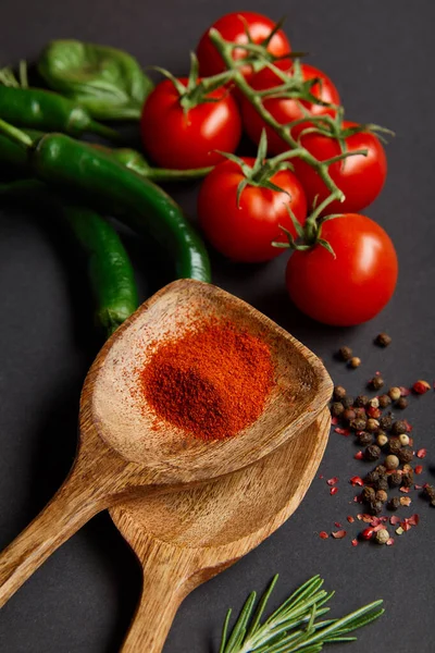 Top view of ripe cherry tomatoes, rosemary, peppercorns, wooden spoons with paprika powder and green chili peppers on black — Stock Photo