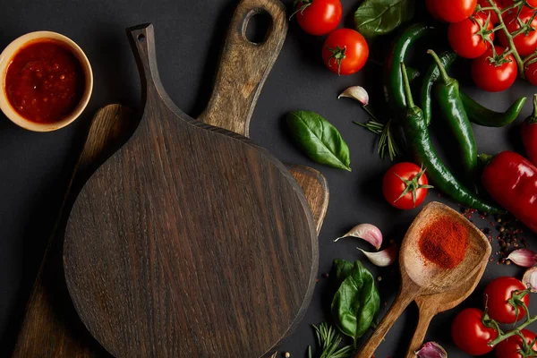 Top view of cherry tomatoes, garlic cloves, tomato sauce, rosemary, peppercorns, and green chili peppers near chopping boards on black — Stock Photo