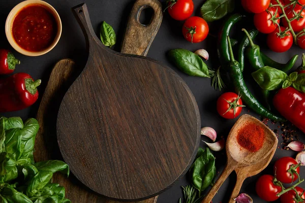 Top view of red cherry tomatoes, tomato sauce, peppercorns, herbs and green chili peppers near cutting boards on black — Stock Photo