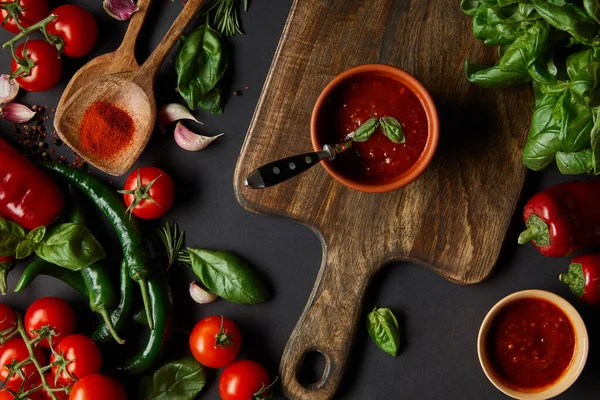Vista superior de tomates rojos cereza, salsa de tomate, granos de pimienta, hierbas y chiles verdes cerca de tabla de cortar en negro - foto de stock