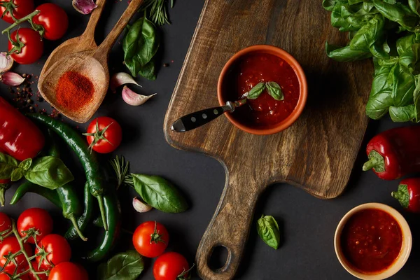 Top view of red cherry tomatoes, tomato sauce, peppercorns, herbs and green chili peppers near cutting board on black — Stock Photo