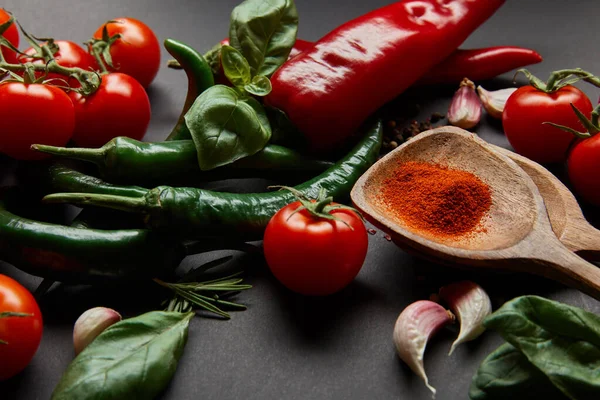 Selective focus of red cherry tomatoes, herbs and chili peppers near spoons with paprika powder on black — Stock Photo