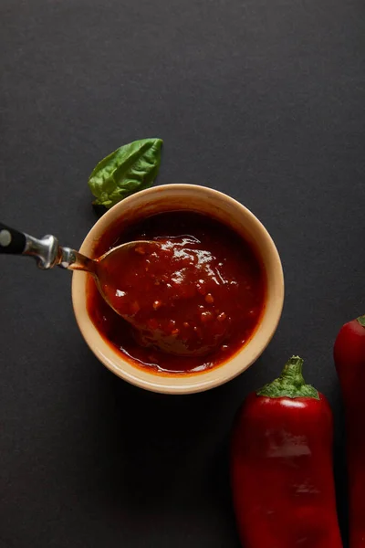 Top view of spoon in bowl with tomato sauce near basil leaf and chili peppers on black — Stock Photo