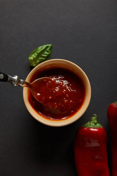 Blick von oben auf Löffel in Schüssel mit Tomatensauce in der Nähe von Basilikumblatt und Chilischoten auf schwarz — Stockfoto