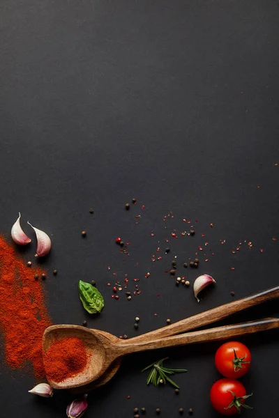 Vista superior de tomates cereja vermelhos, dentes de alho e ervas frescas perto de colheres com páprica em pó em preto — Fotografia de Stock