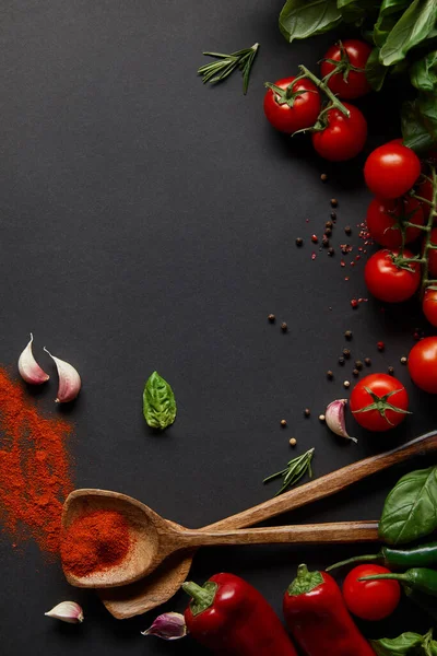 Vista superior de tomates cherry, chiles picantes, dientes de ajo y hierbas frescas cerca de cucharas con pimentón en polvo sobre negro - foto de stock