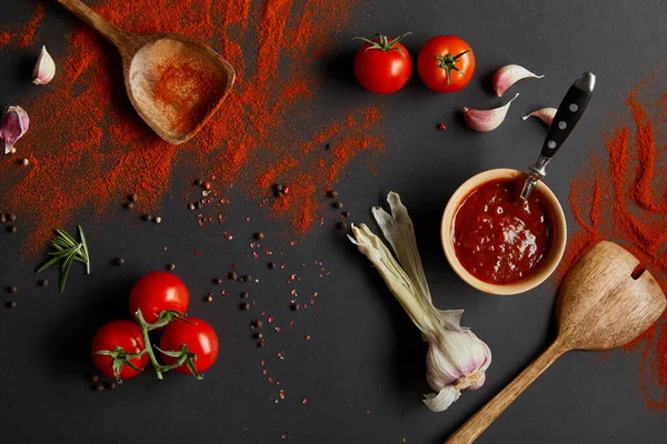 Vista superior de tomates cereja maduros, dentes de alho e ervas frescas perto de colheres de madeira com páprica em pó em preto — Fotografia de Stock