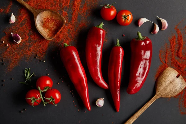 Top view of spicy chili peppers, cherry tomatoes and fresh rosemary near spoons with paprika powder on black — Stock Photo