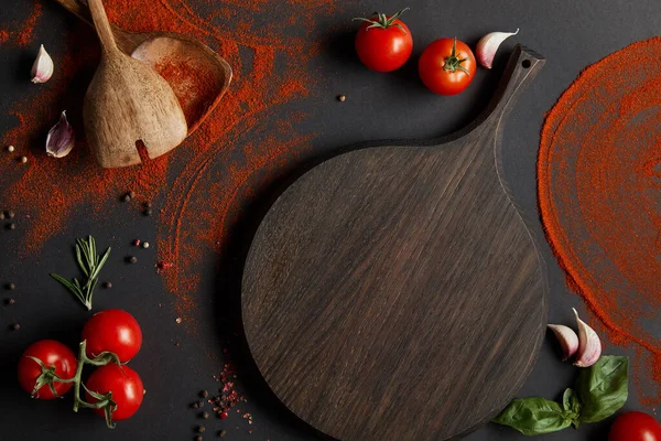 Vista dall'alto di pomodorini rossi ciliegini, tagliere, spicchi d'aglio ed erbe fresche vicino a cucchiai e polvere di paprika su nero — Foto stock