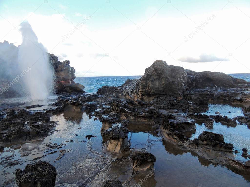 Nakalele Blowhole with water spraying out with ocean and sky in the background, it was created from Pacific Ocean waves hitting the tall rocky cliff coastline that was created from lava on Maui, Hawaii, USA sky reflecting off the still water