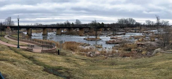 The Big Sioux River flows over rocks in Sioux Falls South Dakota with views of wildlife, ruins, park paths, train track bridge, trees and city in the surrounding area and background