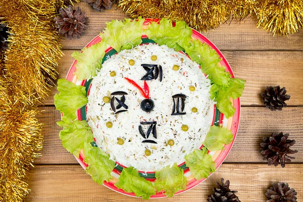 L'insalata di Natale riso olive piselli - concetto Nuovo anno orologio faccia, mezzanotte, marrone sfondo in legno coni di abete rosso fili di lame sul tavolo . Fotografia Stock