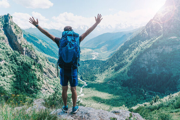a traveler with a backpack on the edge of the cliff raised his hands in the air mountains in the background