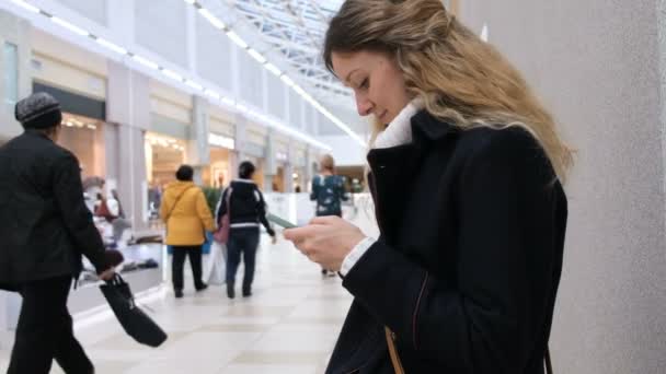 Mladá úspěšná žena kabát a její vlasy získává zprávy do smartphone v obchoďáku. Vypráví o jejich nákup nových přátel. — Stock video