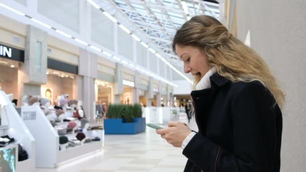 Die junge erfolgreiche Frau im Mantel und mit den langen Haaren bekommt in der Mall Nachrichten aufs Smartphone. erzählt Freunden von ihren Neueinkäufen. — Stockvideo