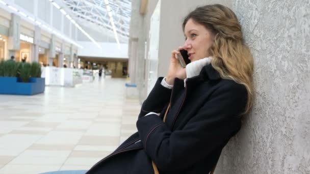 Alışveriş merkezinde bir cep telefonuyla konuşan bir ceket genç çekici kız. Dinlenmeye ve konuşmak için köyde alışveriş yorgun. — Stok video