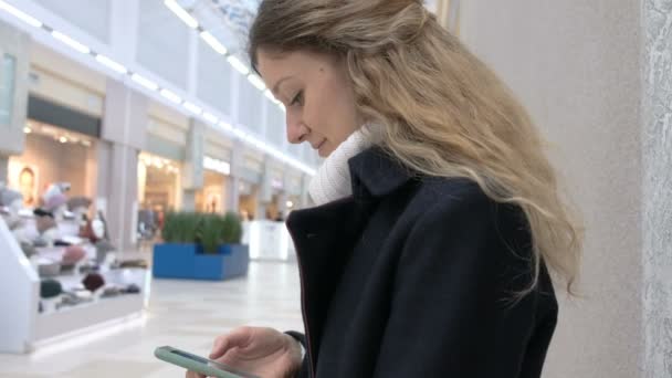 Éxito joven mujer caucásica en abrigo y sus pelos teleconferencia smartphone en el centro comercial. Cuenta a sus amigos sobre sus compras . — Vídeos de Stock