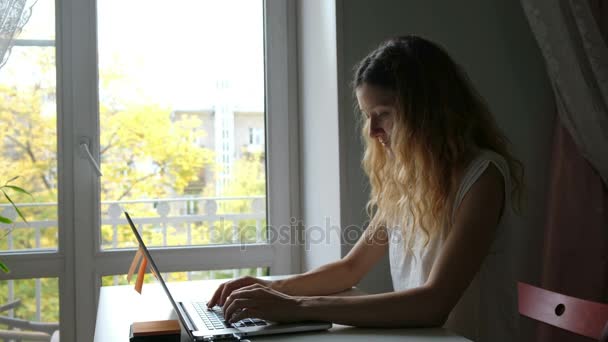 Joven chica caucásica en camiseta blanca trabajando en su casa con su portátil cerca de la ventana. Participó en textos impresos independientes — Vídeos de Stock