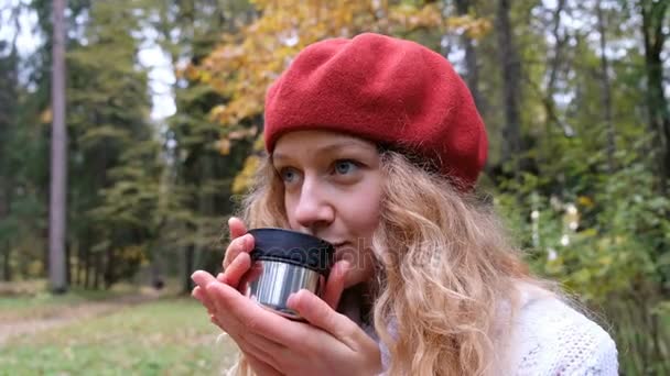 Hermosa joven en una boina roja bebiendo té caliente de un termo y sonriendo, de cerca. Se calienta después de un paseo en el fresco parque de otoño . — Vídeos de Stock