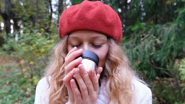 Hermosa joven en una boina roja bebiendo té caliente de un termo y sonriendo, de cerca. Se calienta después de un paseo en el fresco parque de otoño . — Vídeos de Stock