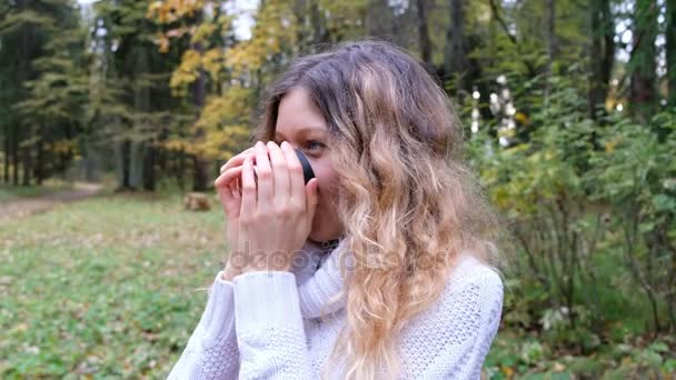 Hermosa joven de ojos azules chica bebiendo té caliente de un termo y sonriendo. Se calienta después de un paseo en el fresco parque de otoño . — Vídeos de Stock