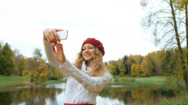 Küçük bir kırmızı hood poz ve modern smartphone bir selfie alarak ve sonbahar Park gülümseyen genç beyaz bir kadın çekici.