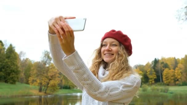 Jovem caucasiana encantadora em um pequeno capuz de equitação vermelho posando e tirando uma selfie em seu smartphone moderno e sorrindo no outono Park . — Vídeo de Stock