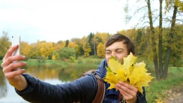 Mladý gay muž blbnout a Hammingova, smích a stal se selfie s podzimní listy na bílém moderní smartphone v parku. — Stock video