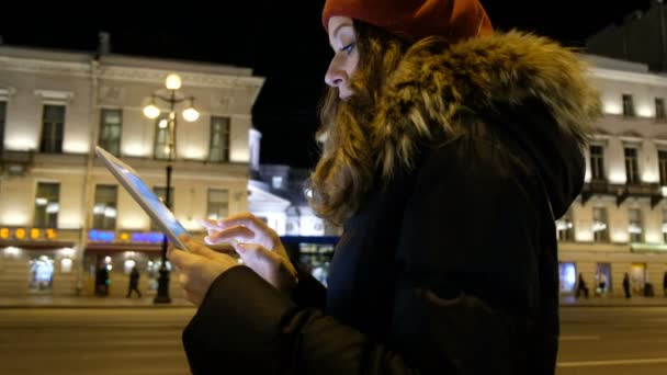 Молодая красивая девушка с планшетом находится на автобусной остановке и ждет автобус в вечернем городе — стоковое видео