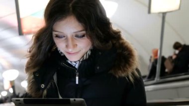 Tablet ekrana bakarak genç çekici beyaz kız ve onun parmaklarıyla tutar, metro dışında yürüyen merdiven üzerinde yükselir. Yakın çekim.
