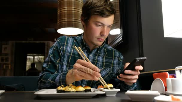 Jovem olha para a tela de um smartphone moderno que está ganhando a mensagem e comendo sushi em um restaurante japonês — Vídeo de Stock