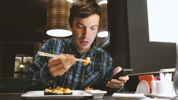 Joven mira a la pantalla de un teléfono inteligente moderno que está ganando el mensaje y comer sushi en un restaurante japonés — Vídeo de stock