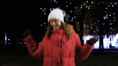 Ağır çekimde, modern smartphone üzerinde kulaklık ile müzik dinlemek ve atlama ve neşe ile kış rojestvensky tatil akşamları dans bir genç çekici beyaz kız.