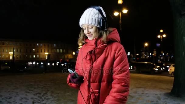 Ελκυστική κοπέλα, να ακούτε μουσική με ακουστικά σε ένα έξυπνο κινητό τηλέφωνο το βράδυ το χειμώνα. — Αρχείο Βίντεο